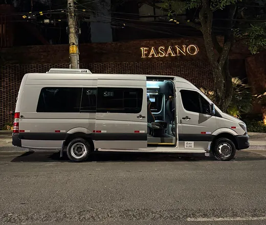 Van executiva prata com a porta lateral aberta diante de um hotel, à noite, pronta para o embarque do grupo